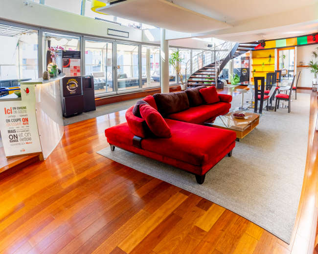 The image shows a modern interior space featuring a large red sectional sofa on a wooden floor, with a staircase, dining area, and large windows allowing natural light.