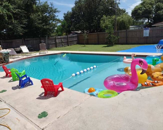 A backyard swimming pool is surrounded by colorful inflatable toys and small plastic chairs, with a wooden fence and grassy area in the background.