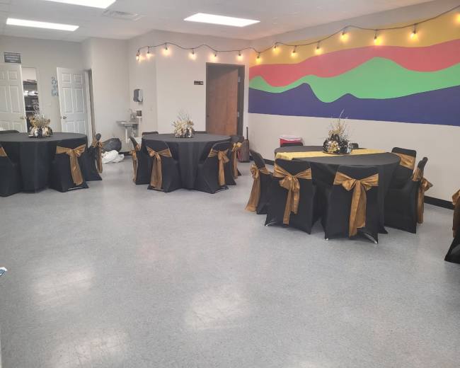 A party setting with several tables dressed in black covers and adorned with gold ribbons, set against a colorful mural on one wall.