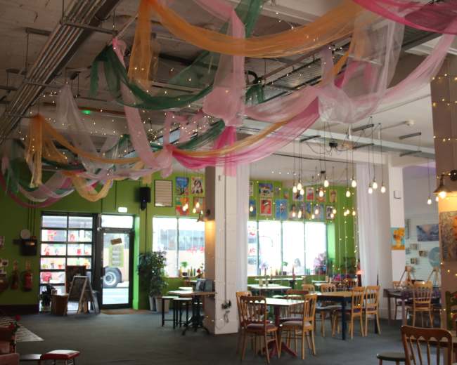 The interior of a vibrant café features colorful drapes hanging from the ceiling, illuminated by string lights, with tables and chairs arranged throughout the space.