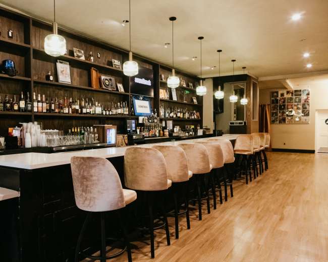 The image shows a well-lit bar with a long countertop, backed by shelves filled with various bottles, and several high-backed chairs positioned in front.