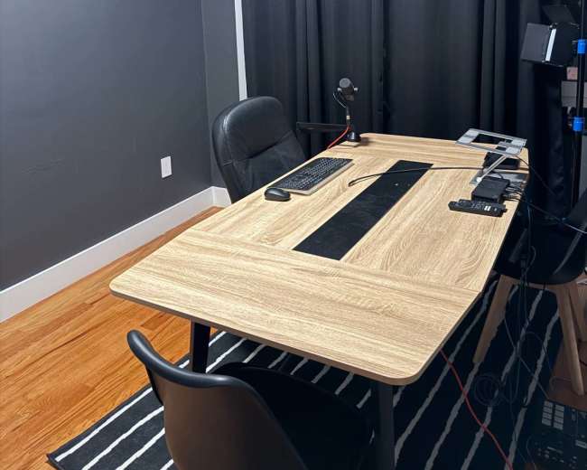 The image depicts a modern home office with a wooden desk, a black chair, and various electronic devices arranged on the desk, all set against dark walls and curtains.