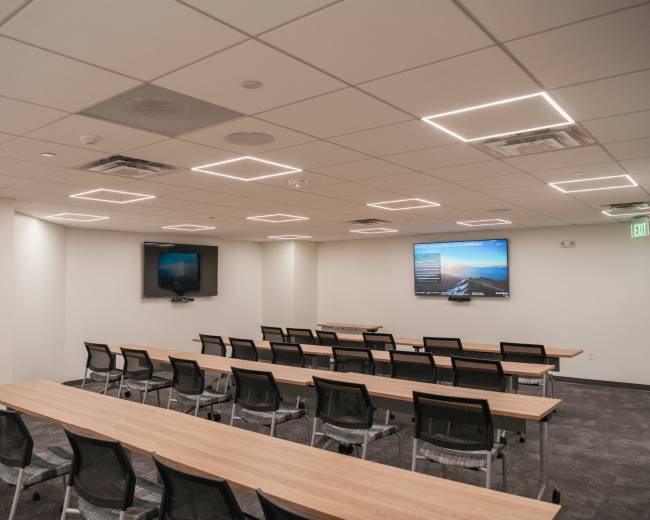 The image shows a modern conference room with rows of tables and chairs, equipped with two screens mounted on the walls.