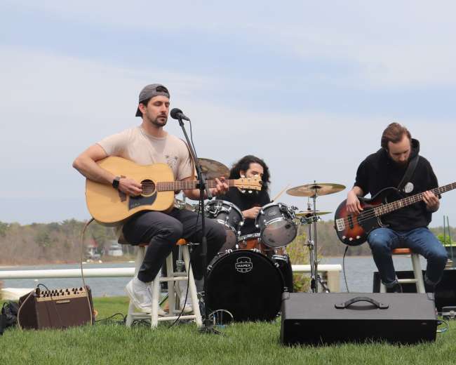 A trio of musicians performs on an outdoor stage by a body of water, with one playing guitar, another on drums, and a bassist.