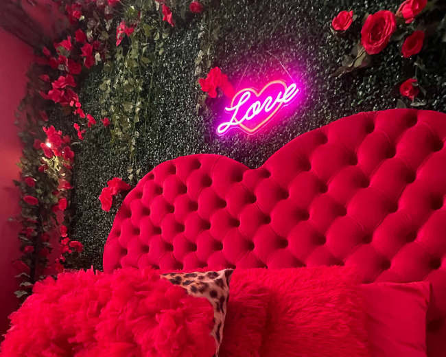 A vibrant bedroom features a pink tufted headboard, red pillows, and a neon sign that reads "Love," surrounded by artificial greenery and floral decor.