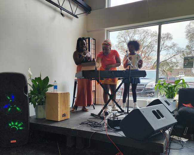 A man and two women perform music on a small stage with a keyboard in a well-lit venue.