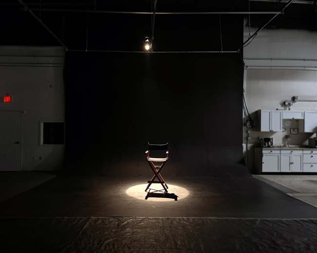 A single director's chair is illuminated by a spotlight on a dark stage with a black backdrop.