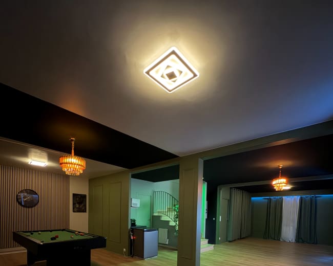 The image shows a spacious room with a pool table, elegant chandeliers, and a staircase leading to another area, illuminated by subtle lighting.