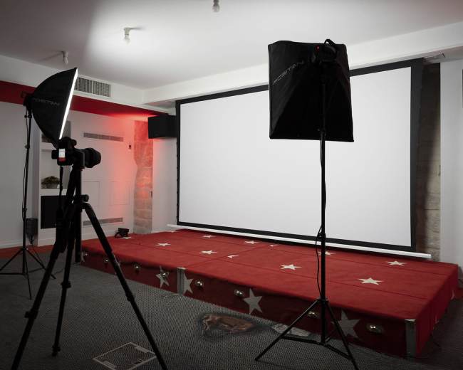 A film screening setup with a large blank projection screen and studio lighting on tripods in a room featuring a red carpet and sound equipment.