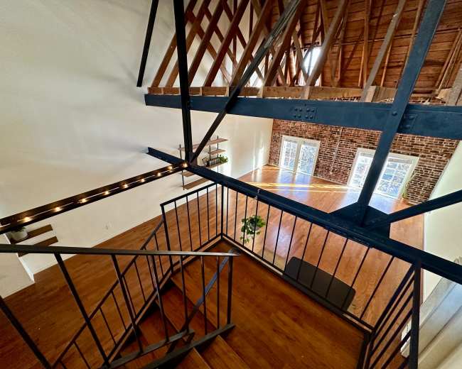 A staircase leads downward into an open space with exposed brick walls and wooden beams, featuring large windows that allow natural light to fill the area.