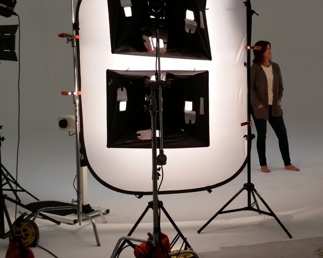 A model stands barefoot in a well-lit photography studio surrounded by multiple lights and cameras.