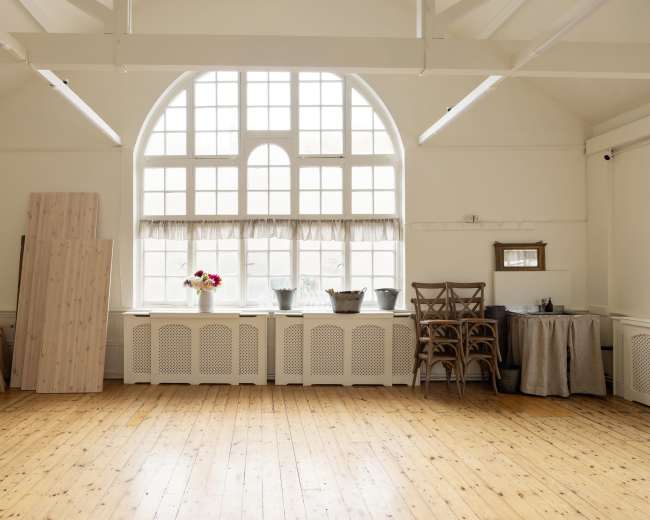 The image shows a spacious room with wooden floors, a large arched window, and various items including planks of wood, a table, and chairs near the walls.