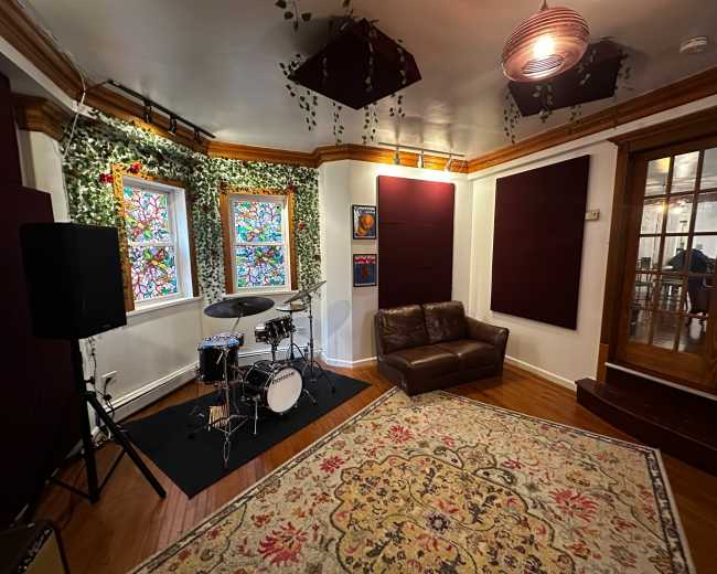 The image shows a music room featuring a small drum set, a brown couch, decorative rugs, colorful stained glass windows, and acoustic panels on the walls.