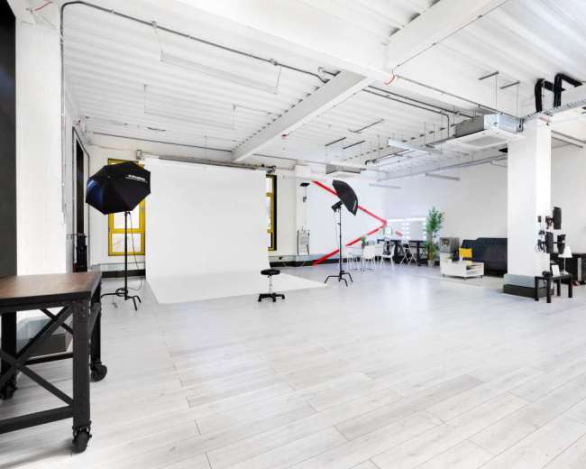 The image shows a spacious, well-lit studio with a plain backdrop, lighting equipment, and various props arranged around the area.