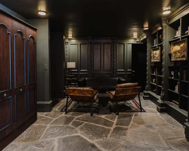 The image shows a dimly lit room featuring a pair of leather chairs and a black sofa, flanked by built-in bookshelves and a large wooden wardrobe.