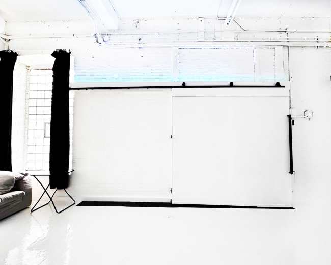 A bright, minimalistic interior with a sliding white door, a sofa, and a small table beside a window with black curtains.