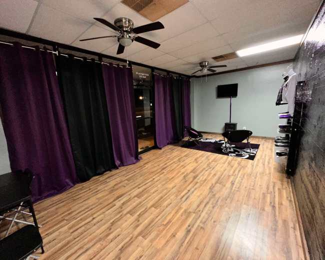 The image shows a vacant room with wooden flooring, purple and black curtains, a wall-mounted TV, and a few pieces of furniture.