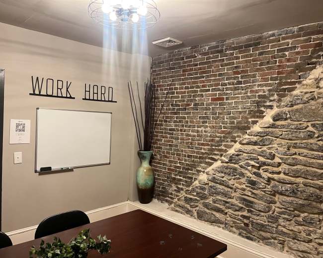 The image shows a meeting room with a wooden table, a whiteboard on the wall, a decorative vase, and an exposed brick feature.
