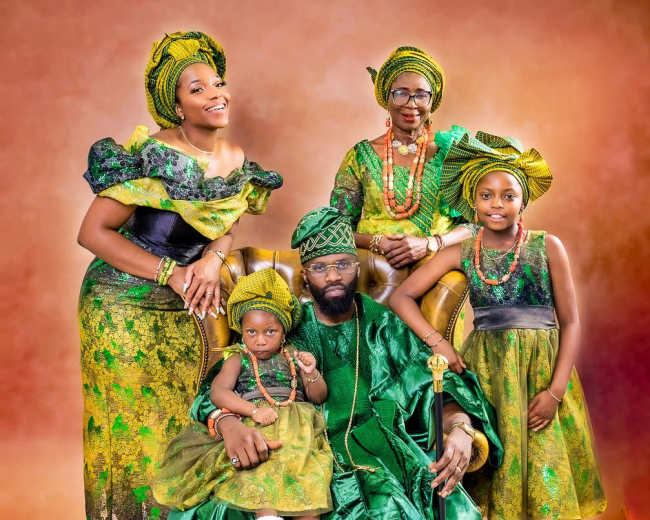 A man in a green traditional outfit sits on a chair surrounded by four women in similar green attire, all posing for a family portrait.