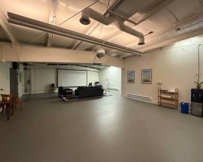 The image shows a spacious, empty room featuring a couch and seating area, a projector setup, and minimalist decor, including two framed pictures on the wall.