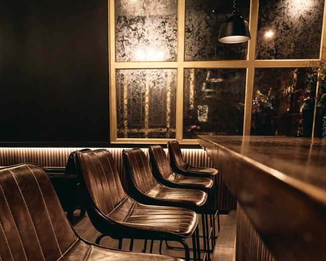 A row of leather chairs in front of a wooden bar, with a large, foggy window behind them.