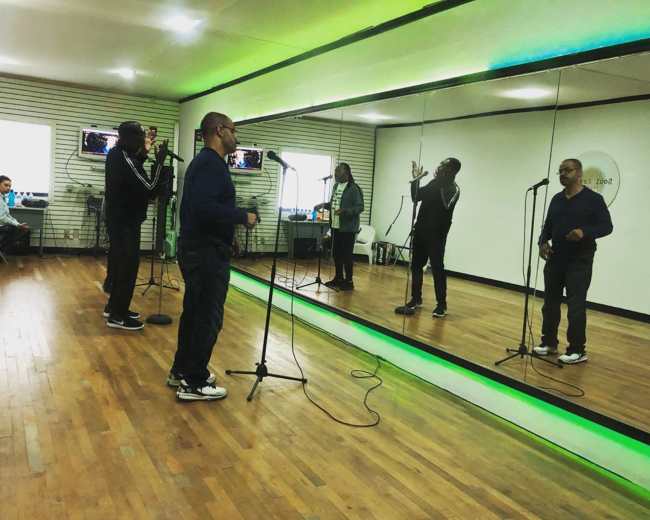 Two men are singing into microphones in a rehearsal studio with mirrors and green lighting.