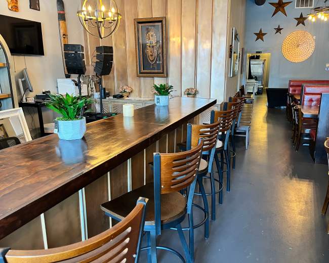 The image shows the interior of a restaurant featuring a wooden bar with high stools, decorated walls, and a pathway leading to dining tables.