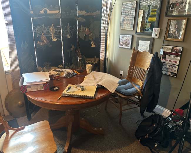 The image shows a cluttered wooden table with an open book, some papers, a coffee cup, and a chair beside it, all set in a small room adorned with framed pictures and a folding screen.