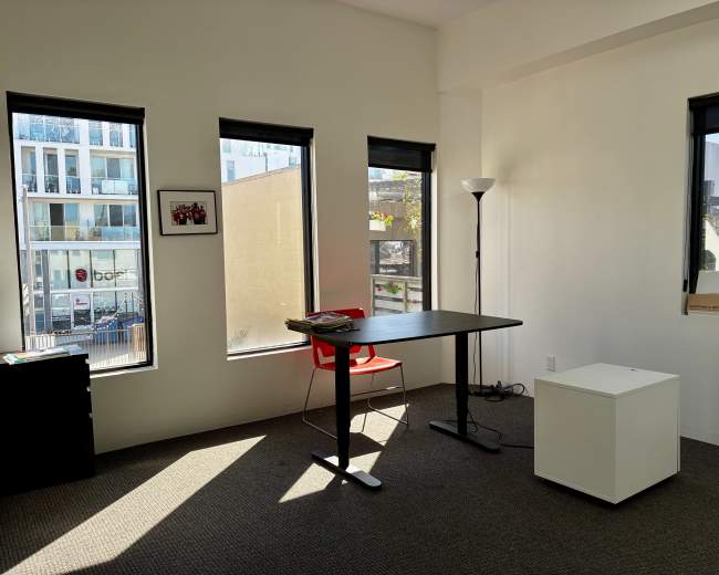 The image shows a minimalist office space with a black table, a red chair, and natural light streaming in through three windows.