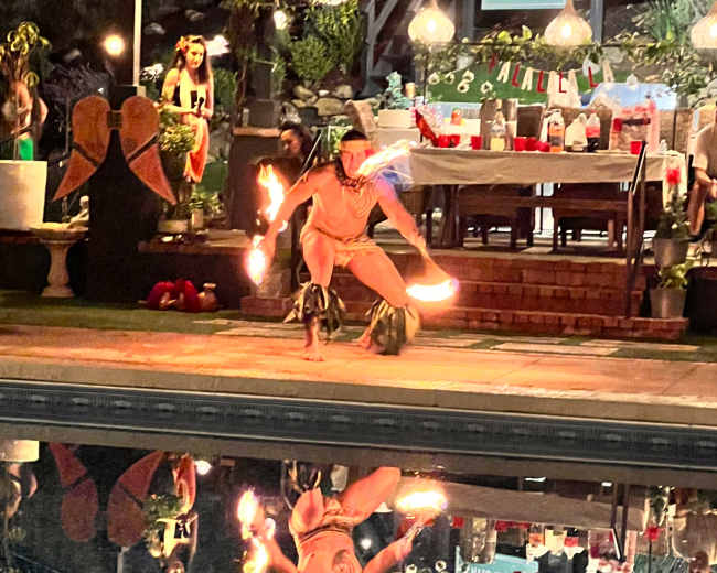 A performer is dancing with fire props by a poolside, reflecting in the water, while a festive gathering is set up in the background.