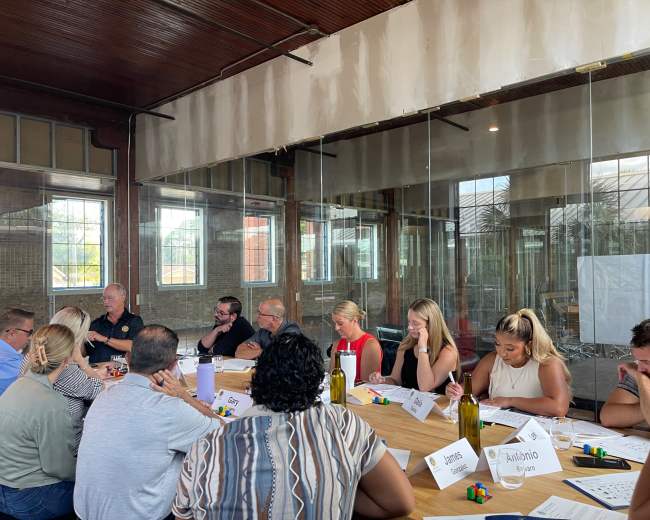 A group of people is seated around a long table in a glass-walled meeting room, engaged in a discussion and taking notes.