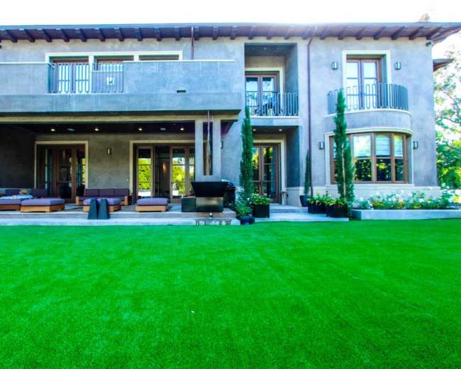 A modern two-story house features large windows, a balcony, and a spacious lawn with artificial grass.