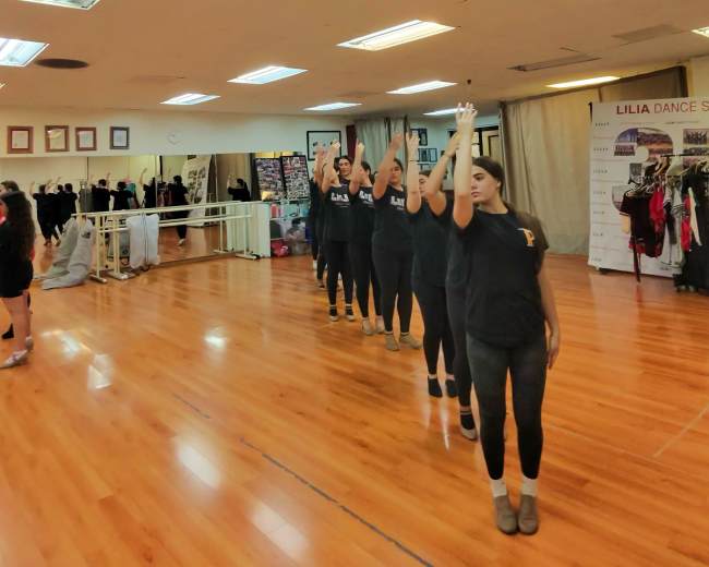 A group of dancers in black shirts practices a synchronized routine in a dance studio with wooden floors and overhead fluorescent lights.