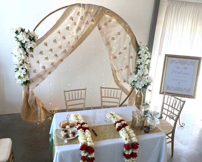A decorative event setup features a gold circular backdrop draped with sheer fabric and adorned with white flowers and fairy lights, accompanied by a welcome sign and a table with a floral arrangement and refreshments.