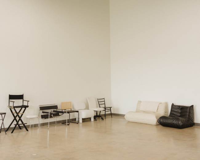 The image shows a spacious, empty room with various styles of seating arranged along the walls.