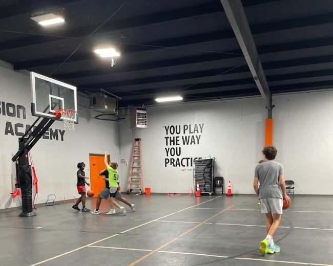 A basketball practice session is taking place in a gym, with players engaged in drills near a hoop and a wall featuring motivational text.