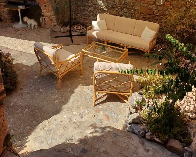 The image shows a cozy outdoor seating area with a large umbrella, surrounded by stone walls, featuring a rattan sofa, chairs, and a coffee table.