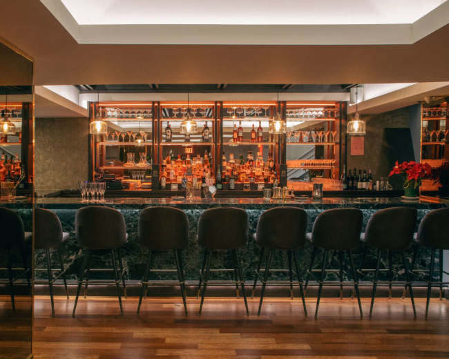 The image shows a modern bar with a marble counter, stylish seating, and shelves lined with various bottles of liquor.