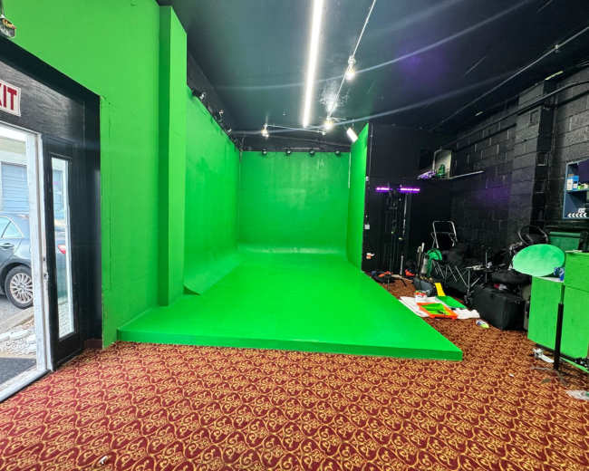 The image shows a room with a green screen setup along one wall, and various equipment and props scattered around a patterned carpet.