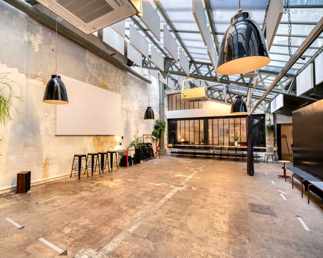 The image shows a spacious, industrial-style interior with concrete floors, large pendant lights, and a minimalist setting featuring a projection screen and seating area.