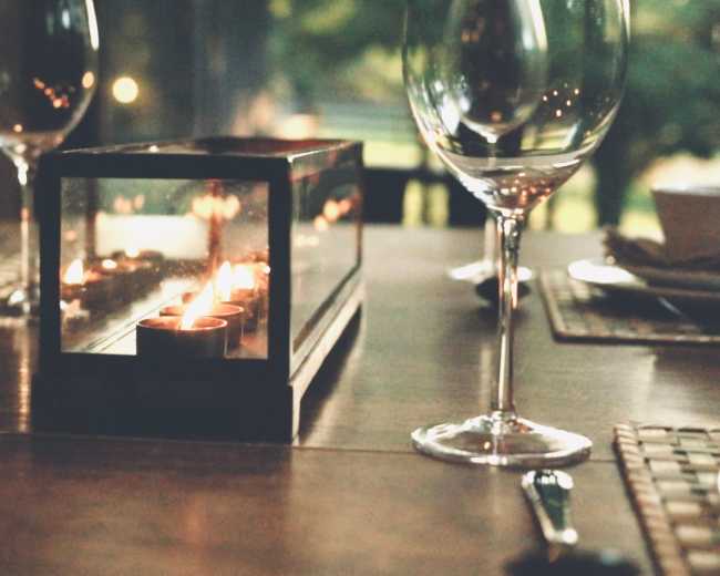 A dining table is set with a glass candle holder and empty wine glasses, surrounded by a natural outdoor setting.