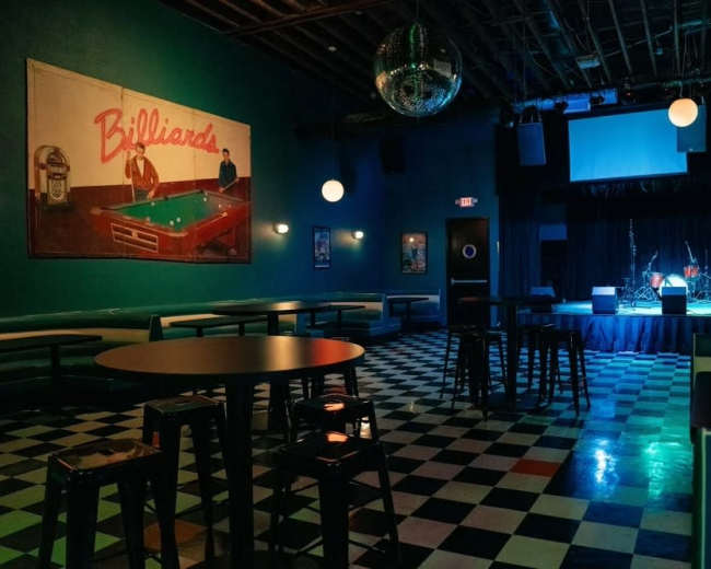 The image shows an empty venue with checkered flooring, a stage at the front, and a mural of a billiards scene on the wall.
