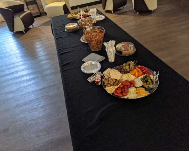 A long table is set up with various food items, including a cheese platter, crackers, fruits, and snacks, under low lighting.