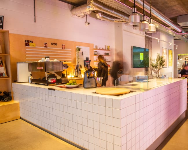 The image shows a modern café with a tiled serving counter, a coffee machine, and a seating area illuminated by natural light.