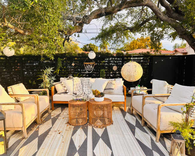 The image shows a cozy outdoor seating area featuring light-colored sofas and chairs arranged around woven coffee tables, with greenery and hanging decorations overhead.