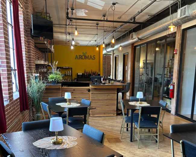 The image shows a modern dining area with wooden tables and chairs, illuminated by pendant lights, leading to a well-equipped bar in the background.