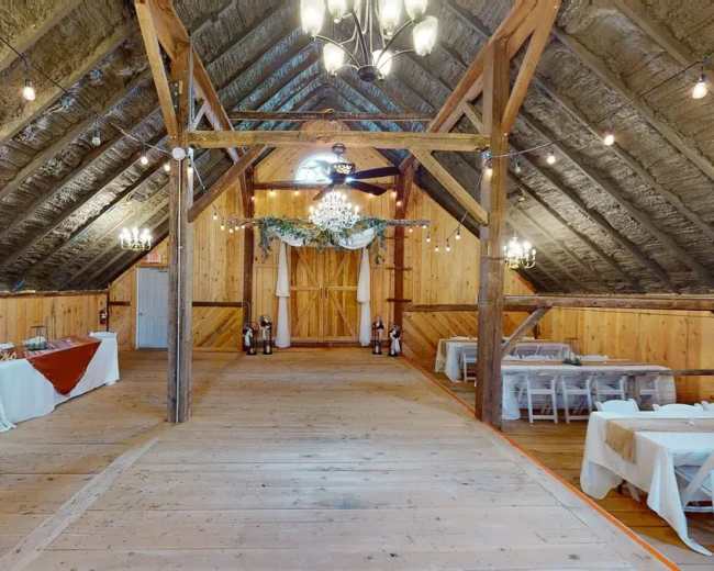 The image shows the interior of a rustic barn venue with wooden walls, a wooden floor, and tables arranged for an event.