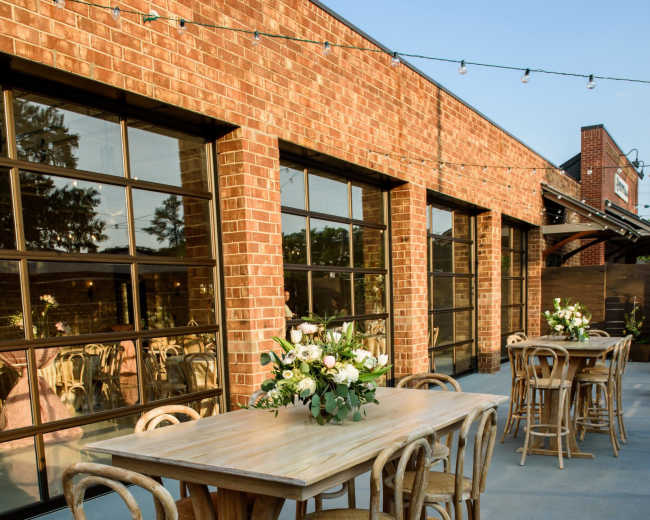 The image shows a patio area with wooden tables and chairs, adorned with floral centerpieces, next to a brick building with large windows.