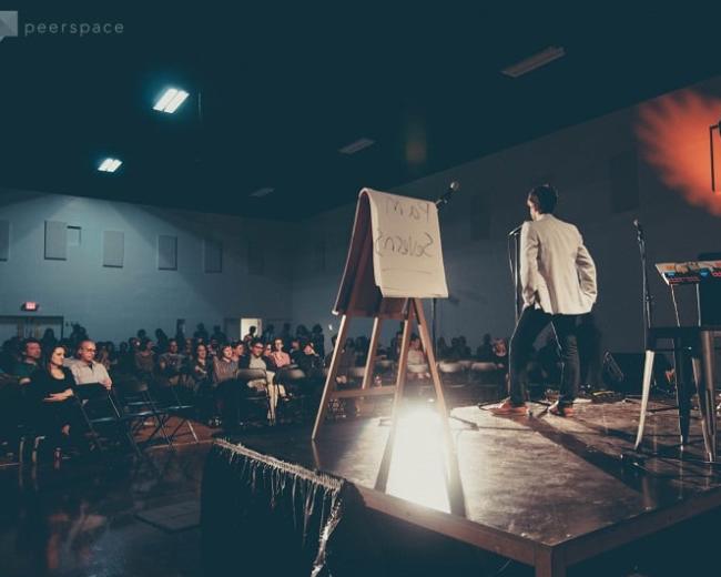A speaker stands on stage in front of an audience, with a blank presentation board and equipment nearby.