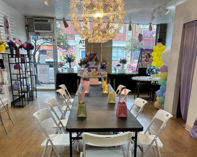 A table with white folding chairs is set up in a brightly lit room adorned with colorful decorations, including flowers and balloons.